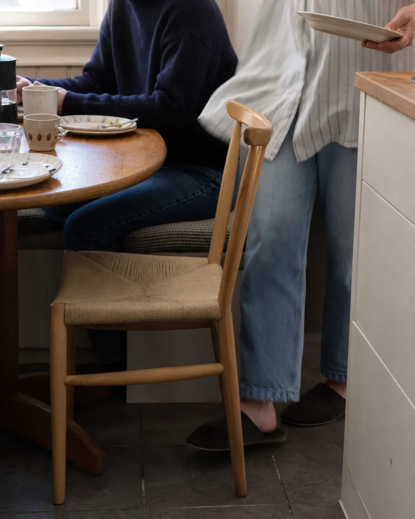 One person seated at a breakfast table while another passes behind a pulled-out chair toward the kitchen.
