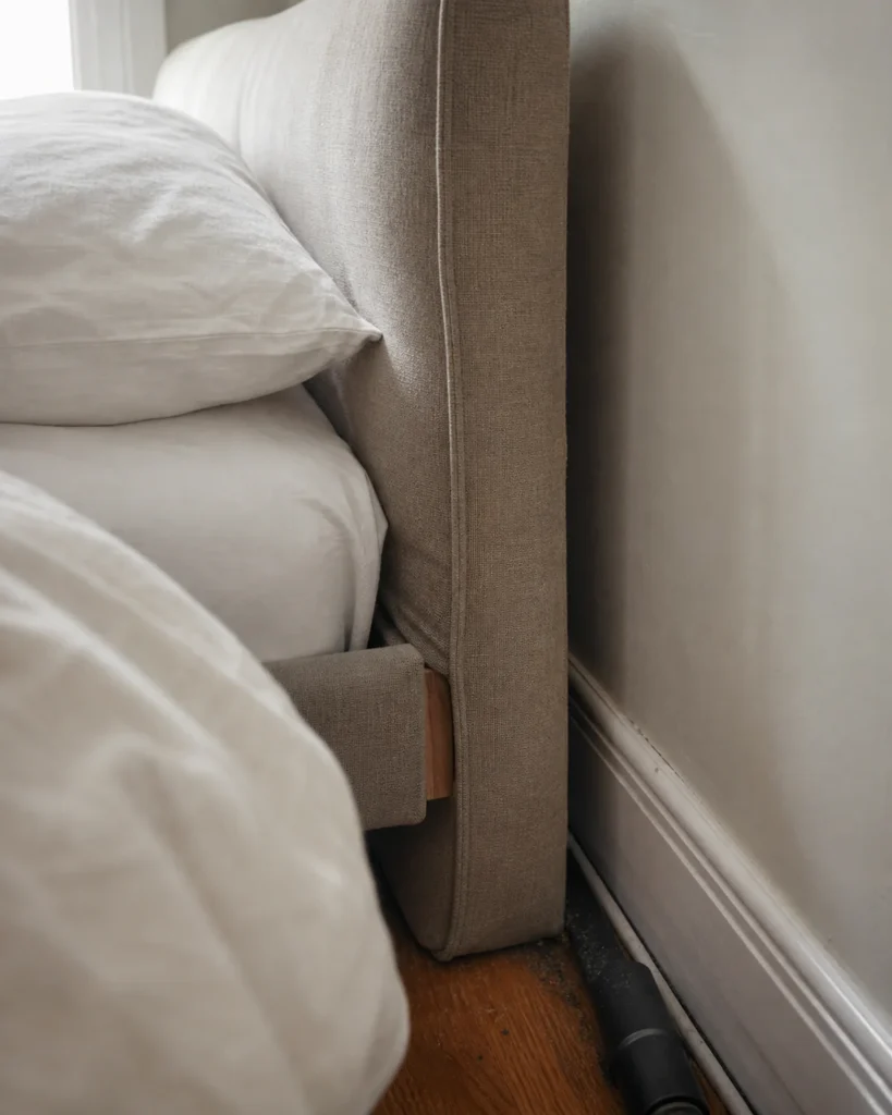 Side profile of an upholstered headboard showing its fabric seam, wall gap, skirting and a vacuum crevice tool.