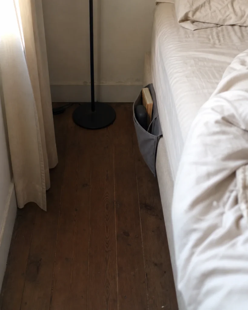Narrow bedside gap using a floor lamp and purpose-made fabric bed pocket instead of a nightstand.