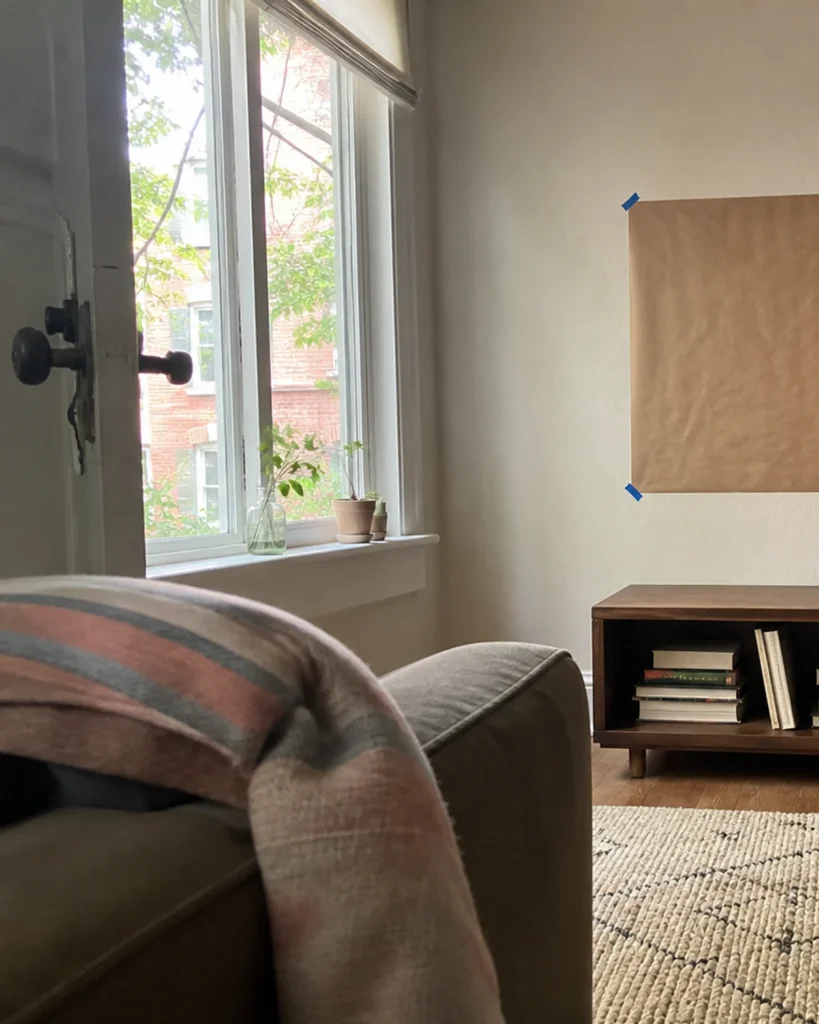 View from the end of a sofa toward a paper television outline while daylight from the opposite window crosses the proposed screen position.